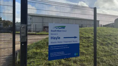 A fence surrounds an industrial looking building. A sign outside says South west water - Hayle treatment works.