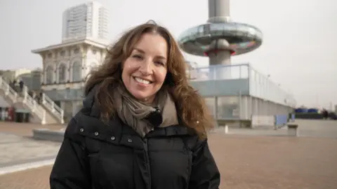 A brunette woman in a black puffa jacket smiles at the camera in front of Brighton's i360 building.