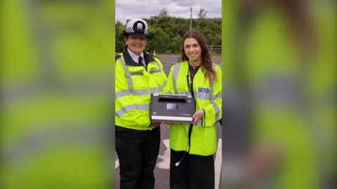 Two women, one in Police uniform and the other in a Police high-viz hold a breathalyser and smile at the camera.