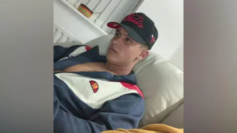 Family Handout A person wearing a black cap and a zip‑up jacket is sitting on a light‑coloured sofa indoors.
