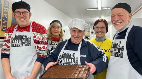 Andrew Turner/BBC Five people stand inside a kitchen. Four are wearing aprons and bakers' hats. The woman in the middle is holding a tray of freshly-baked items, possibly gingerbread. All are smiling.