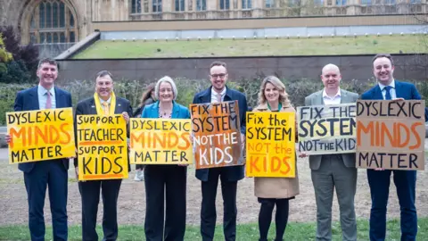 Livvi Grant A group of people are standing in a row holding signs outside parliament. Adam Dance is in the middle holding a sign that reads fix the system not the kids.