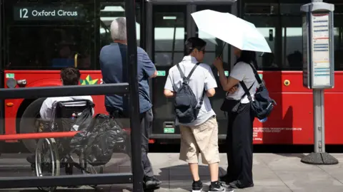 Toby Shepheard/AFP/Getty Images A picture showing people at a bus stop in London with the number 12 bus to Oxford Circus at the stand. From left to right there is a man in a white shirt who is a wheelchair user, a man in a blue shirt and black trousers leaning on the cover and two people under a white umbrella - one is a man in a light coloured shirt and brown trousers, and a woman in a white shirt and black trousers also wearing a backpack.