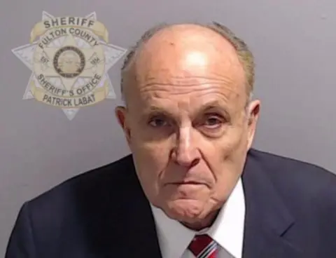 Fulton County Sheriff's Office A police mugshot showing Rudy Giuliani in a blue suit and red and blue striped tie which appears to be based on the Stars and Stripes