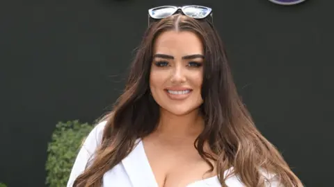 Getty Images Amy Christophers, in a white dress, smiling at the camera. Her hair is down and she is wearing sunglasses on her head