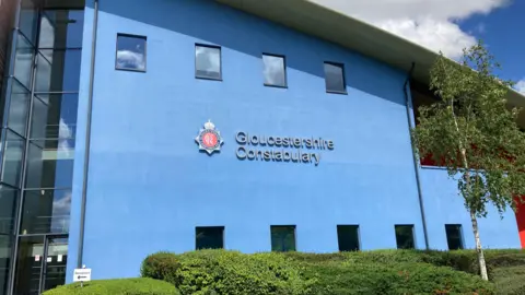 The front of the Gloucestershire Constabulary headquarters. There is a large blue painted wall with small windows on it and a sloping roof. There are bushes and trees outside. There are white clouds and patches of blue in the sky.