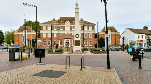 Thurrock Council A landscape image of The Old Courthouse from Grays High Street. In front of the large building is a small cenotaph. There are also black bollards and a lamppost in the image and a delivery driver on a moped. 
