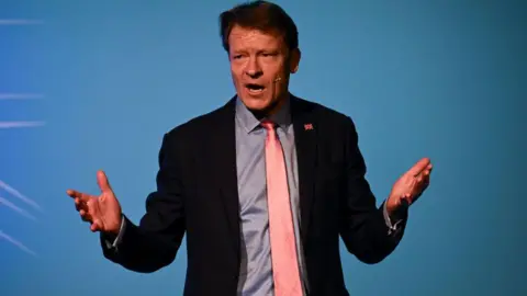 Getty Images Richard Tice, standing on a stage in front of a light blue background. He is mid speech with both arms outstretched. He is wearing a dark suit, blue shirt and pink tie.