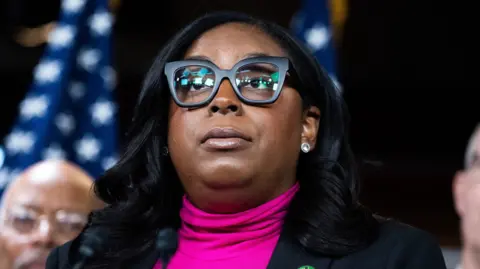 LaMonica McIver wears a bright pink jumper, a black jacket and black glasses as she looks up.