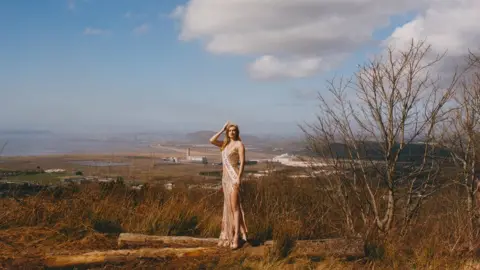Roo Lewis A woman with a Miss Wales sash stands on a hill with the steel works in the backdrop