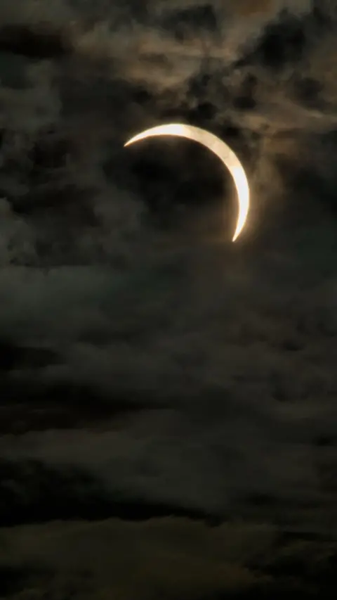 A solar eclipse hangs over the Cumbrian landscape, with the darkened Sun surrounded by a glowing corona above rolling hills and a dramatic sky.
