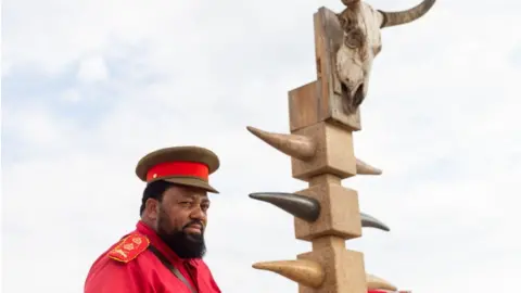 Getty Images A high-ranking chief in traditional Herero clothes stands next to a monument in honour of the OvaHerero/OvaMbanderu and Nama people that were victims of the genocide by German colonial forces at the beginning of the 20th century stands at the Swakopmund Concentration Camp Memorial, in Swakopmund, Namibia, on March 27, 2019