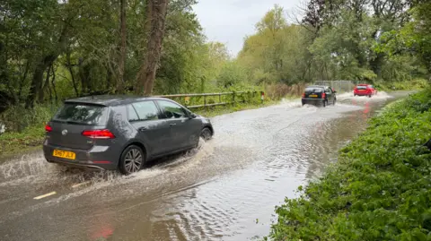 BBC Three cars driving through floodwater on a country road. The water looks to be several inches deep. Either side of the road is greenery, bushes and trees.