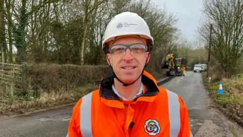 Ollie Conopo/BBC Man in hard hat and orange hi-viz stands on a road with the pothole machine in the background