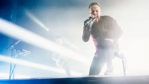 Getty Images Sam Carter on stage. He's screaming into a microphone, with one leg elevated on a stage monitor in front of him. Bright white stage lights are flooding the space behind him.