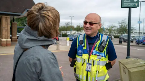 Northamptonshire Police A police officer wearing a high-vis gilet speaking to a person wearing a grey hoodie, who has their back turned to the camera. He appears to be in a supermarket car park.