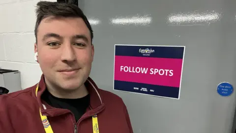 Luke Dudley Luke wearing a Eurovision lanyard and standing by a door with a sign saying 'follow spots'