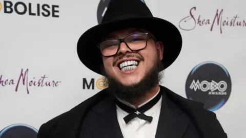 PA Media Potter Payper at the Mobo Awards in Sheffield. Potter wears a black fedora hat, a black blazer and a thin black bow tie over a white shirt. He smiles at the camera, revealing a gem-encrusted grill. He wears black rimmed glasses and has a dark brown beard and moustache