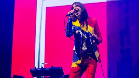 BBC A man wearing a multi coloured jacket, shirt and trousers, sings into a microphone while performing on stage.