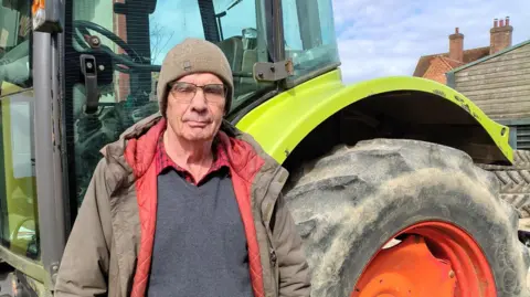 Mike Pinard, wearing a jacket and hat, stands beside a large green tractor with a red wheel, on a farmyard with buildings in the background.