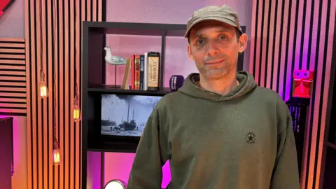 A man wears a hoody and a baseball cap, standing in what looks like a cool barber's shop 