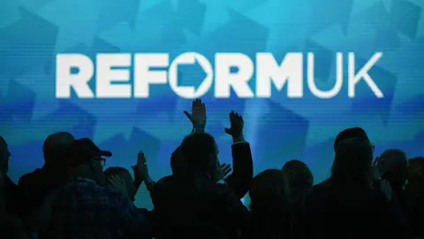 A Reform UK slogan projected on a wall with crowds pictured below in the dark.