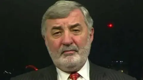 Lord Alderdice has grey hair parted to the side with a grey beard and is wearing a pin striped dark suit
