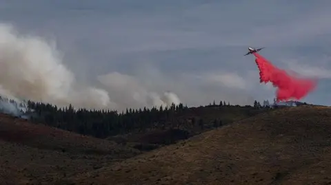 A plane releases a red chemical substance on land near a wildfire in attempt to curb the spread.