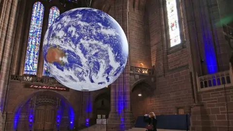 In the centre of the image is a giant scuplture of the earth called Gaia, covered in satellite imagery of the planet. It's being suspended from the ceiling. It's being suspended in Liverpool Cathedral - and globe is surrounded by stained glass window, tall brick walls, and a staircase to the right.