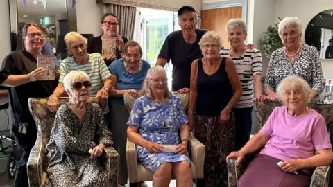 Langham Manor A group of people are sitting down and standing up. They are all looking towards the camera. Some of them are holding books which are written by Larry Lamb.