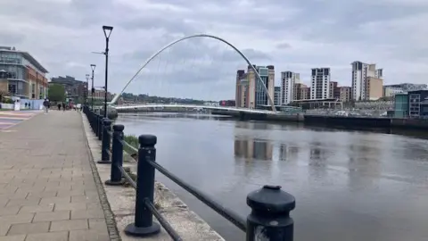 BBC/Luke Walton The Millennium Bridge on an overcast day. The arched bridge is reflected in the still water and can be seen in the distance beside the quayside and black posts and a fence at the water's edge. The Baltic arts centre can be seen on the other side of the water. 