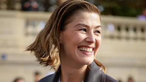 Close - up head shot of Roasamund Pike with bob length light brown hair smiling and looking to the right in the sun in Paris. She is wearing a dark grey jacket
