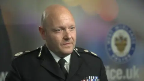 A bald man wearing a black uniform looks to the right of the frame. He is in front of a blurred background with the badge of West Midlands Police in frame.