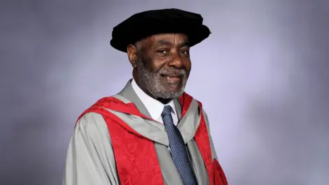 UWE Bristol Guy Bailey OBE. He is pictured wearing a graduation cap and gown. He is smiling at the camera. 