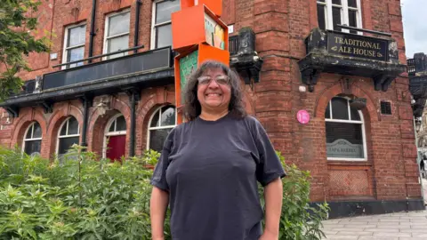 Simon Thake An Asian woman with curly dark hair and glasses stands proudly in-front of a red brick building