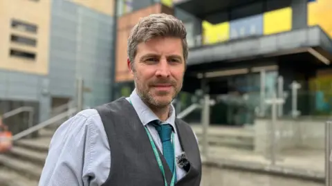 Councillor Phil Alford in waistcoat and tie standing outside the council headquarters, looking at the camera.