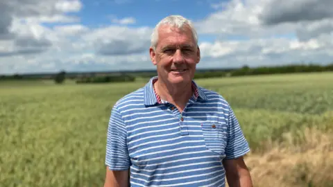 Kevin Shapland, wearing a blue and white striped shirt standing in front of a field.