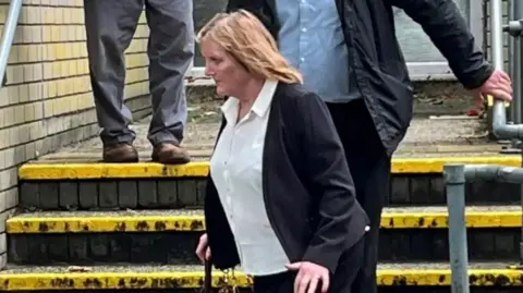 Bridget Curtis outside court. She is wearing a dark blazer with a white shirt and has shoulder length blonde hair. She has a walking stick in one hand and can be seen in front of a set of three steps. 