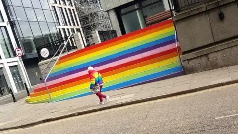 Neil Goodbrand Lady in rainbow jacket in front of rainbow
