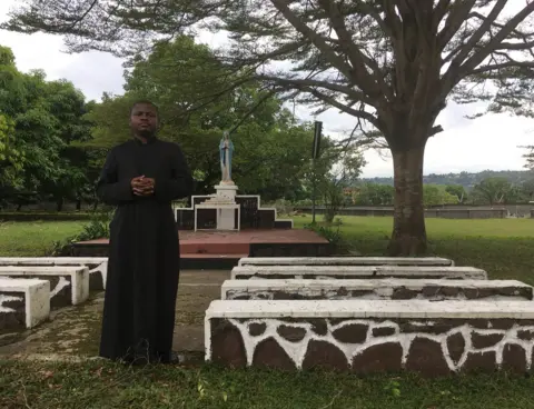 Alice Doyard/BBC Father Joseph Musubao wears his black cassock and stands close to an outdoor altar and pews