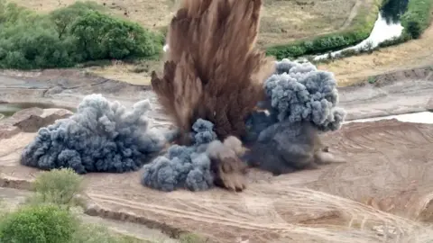Smoke and dirt explode into the air on a site filled with sand and grass.