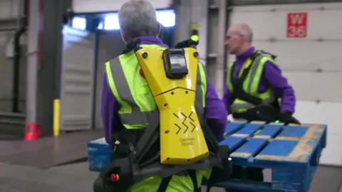 Two warehouse workers wear a yellow exoskeleton to help them carry a blue wooden palette.