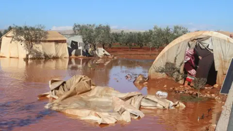 IYD/CARE A woman stands in her flooded tent in Idlib province, Syria