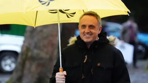 Alex Cole-Hamilton, who has short grey hair, smiles while standing under a yellow Lib Dem branded umbrella 
