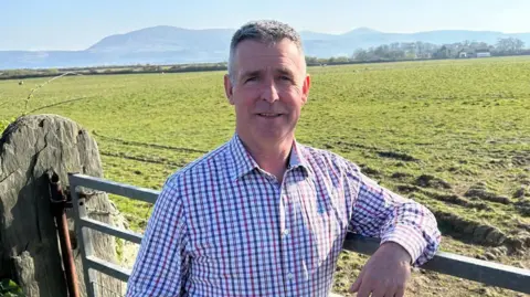John Teare John Teare has short grey hair, and wears a blue and red checked shirt. He leans on a farm gate near a green field.