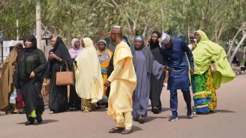 A group of men and women, dressed in northern Nigerian Muslim attire, walk on the road outside the home of Muhammadu Buhari. There are green trees behind them.