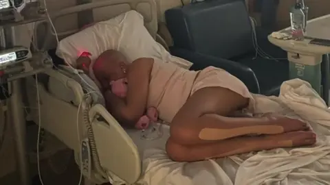 Lyra Cassell Ms Cassell, who has lost her hair during treatment, is lying on her side in a hospital bed and is cuddling a pink teddy bear. She is surrounded by medical equipment. 