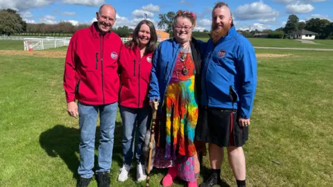 Bargain Hunt contestants - Carl, Kim, Kirsty and Simon. Carl and Kim are wearing red fleeces and Kirsty and Simon are wearing blue fleeces. All four of them are standing in a line, looking at the camera and smiling. 