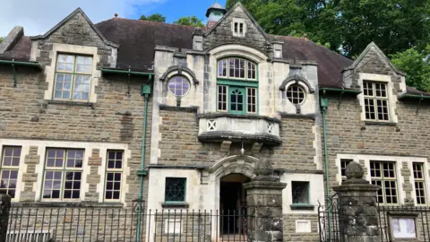 Oakdale Miners' Institute as it stands in St Fagans National Museum of History. It is a large, two-storey building made of large stone walls. It has cream and green window frames. In the foreground is a stone wall with iron railings and behind the building are some large trees.