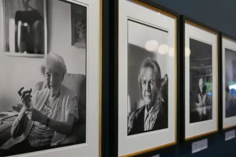 Mike Stone A side-angle shot of several framed black and white photographic portraits of elderly individuals hanging in a row on a gallery wall.